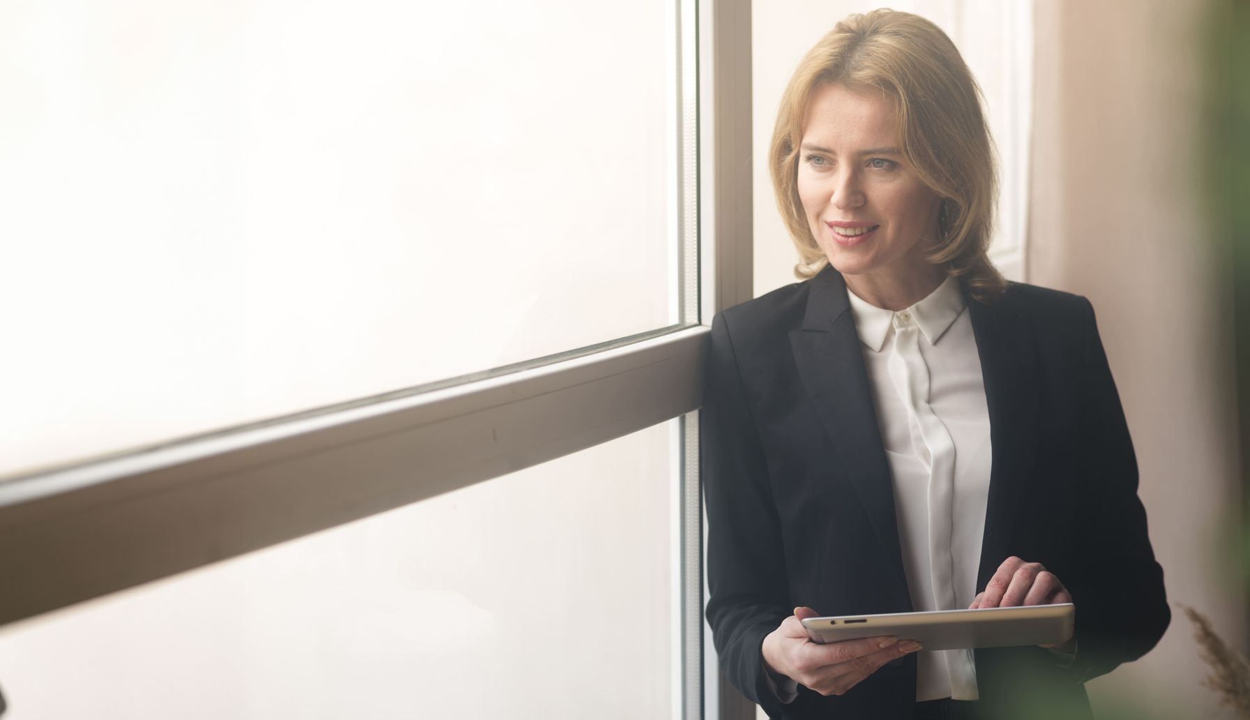 A confident businesswoman in a black suit holding a tablet, standing by a window with natural light.